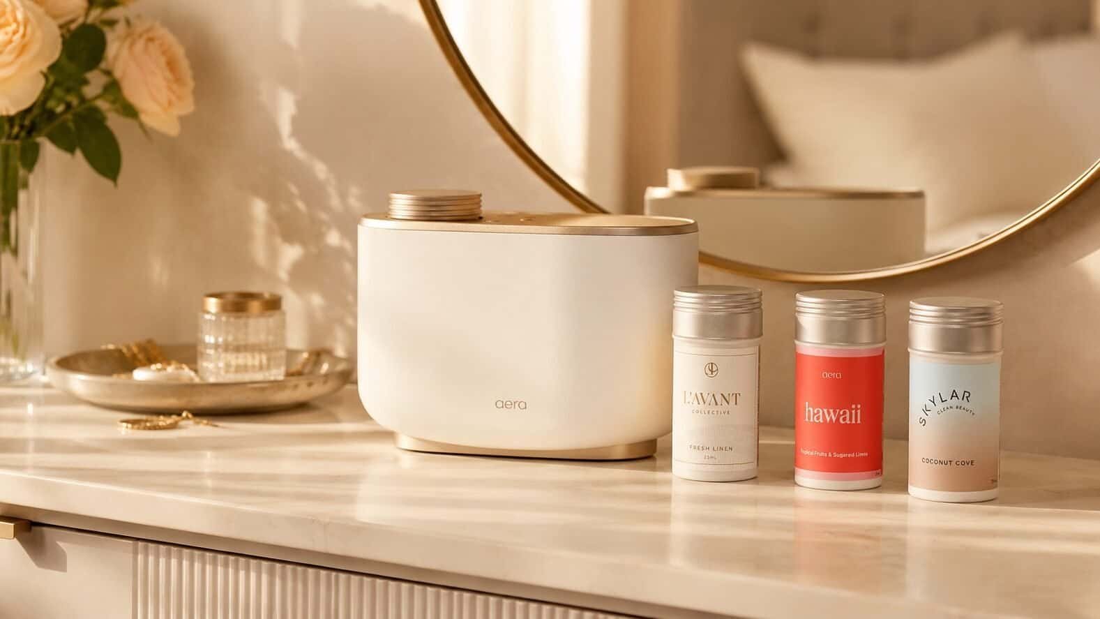 Aera Review image featuring the Aera Diffuser on a bedroom vanity with three fragrance capsules, soft warm lighting, flowers, and elegant bedroom decor.