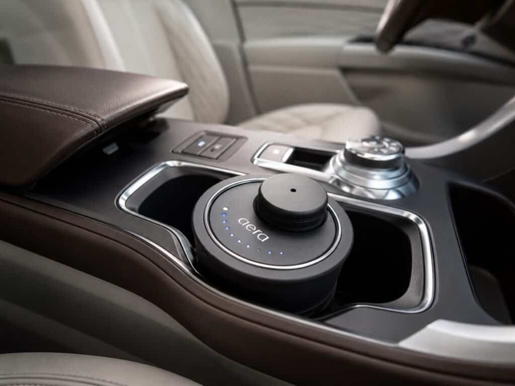 Aera Go diffuser placed in a car cup holder, shown in close-up inside a luxury beige leather car interior with a dark center console.