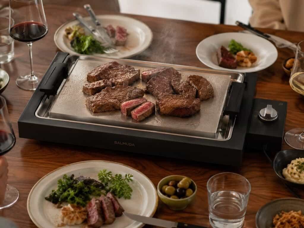 Balmuda The Teppanyaki electric grill on a dining table with steak and vegetables cooking on the steel surface