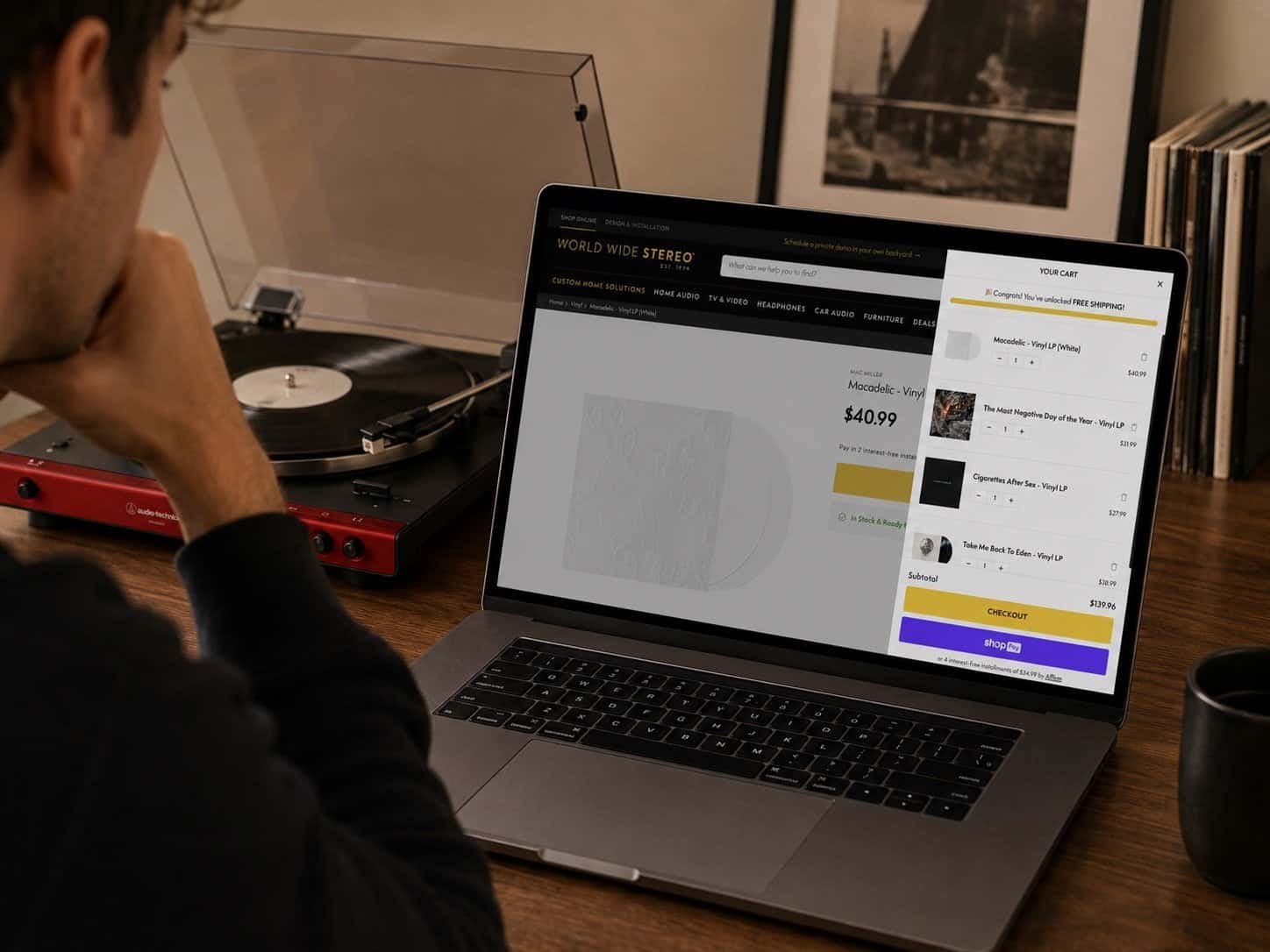 Person browsing vinyl records in a Worldwide Stereo shopping cart on a laptop beside a turntable.